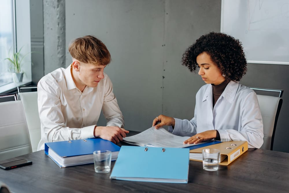 Two reviewers at a boardroom table turning the pages of a long document, with ring binders of paperwork stacked in front of them