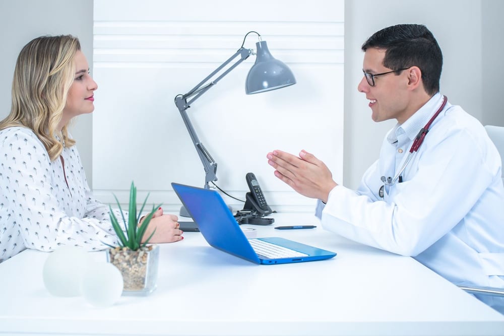 A practitioner in a white coat talking with a seated client across a desk in a bright consulting room