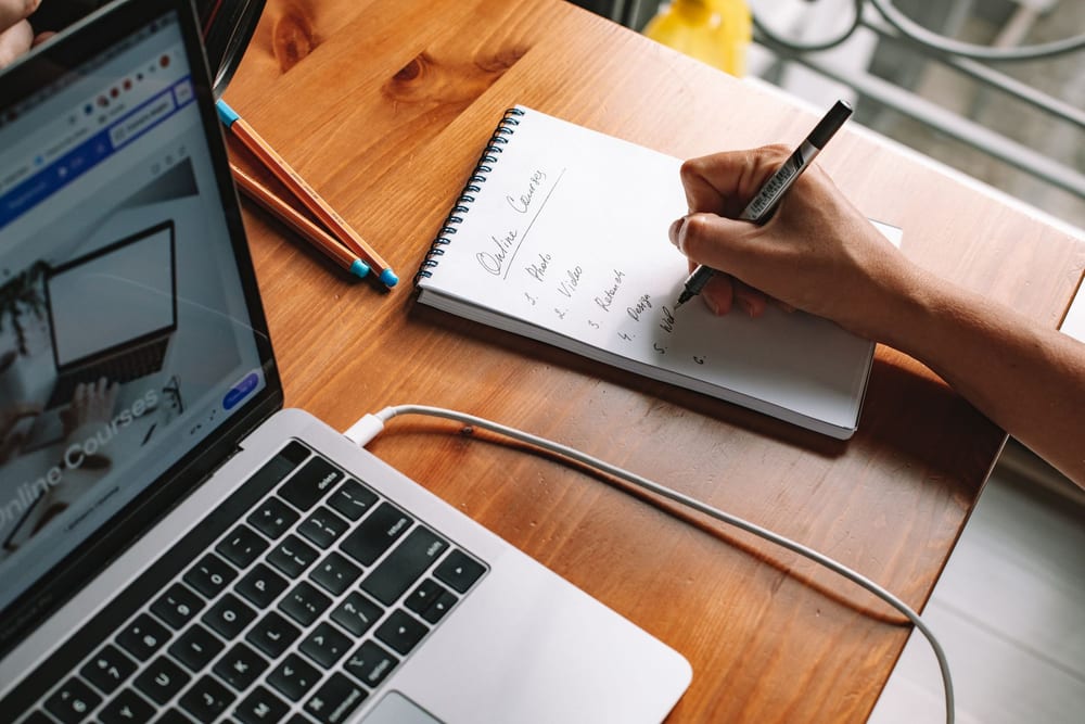 A hand writing a numbered list of online courses into a spiral notebook beside an open laptop showing a course catalogue