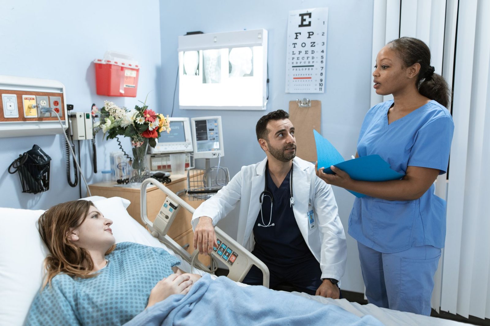 A clinician in a white coat leaning at the rail of a hospital bed while a colleague in blue scrubs stands opposite holding an open patient chart, the kind of in-person clinical site where dietetics supervised practice is completed