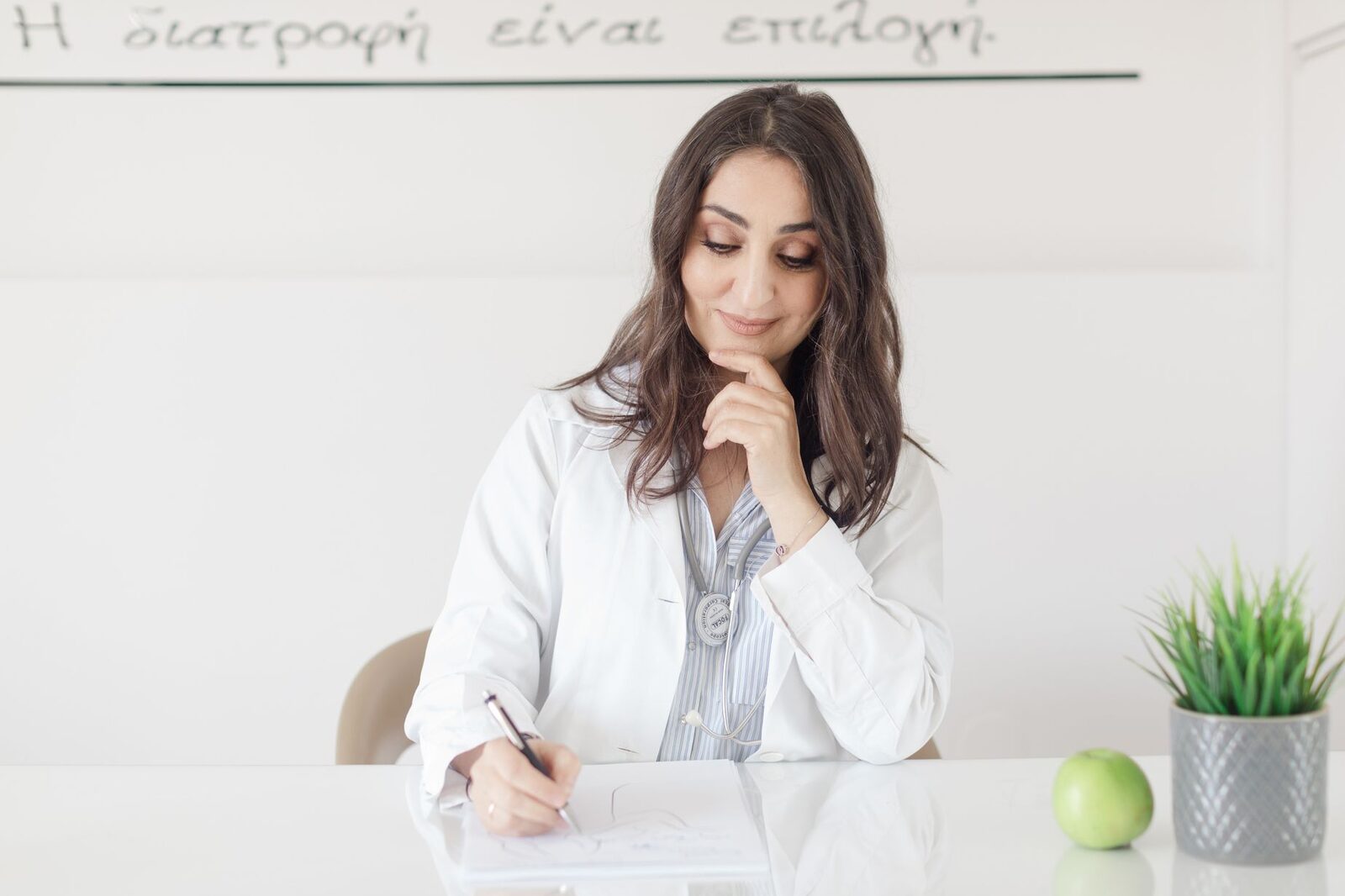A nutrition professional in a white coat writing notes at a clean desk, with a green apple and a potted plant beside her
