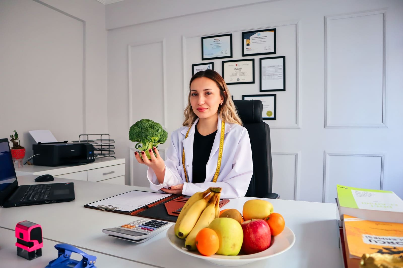 A nutritionist in a white coat seated at a desk holding a head of broccoli, with a bowl of fruit in front of her and framed certificates on the wall behind