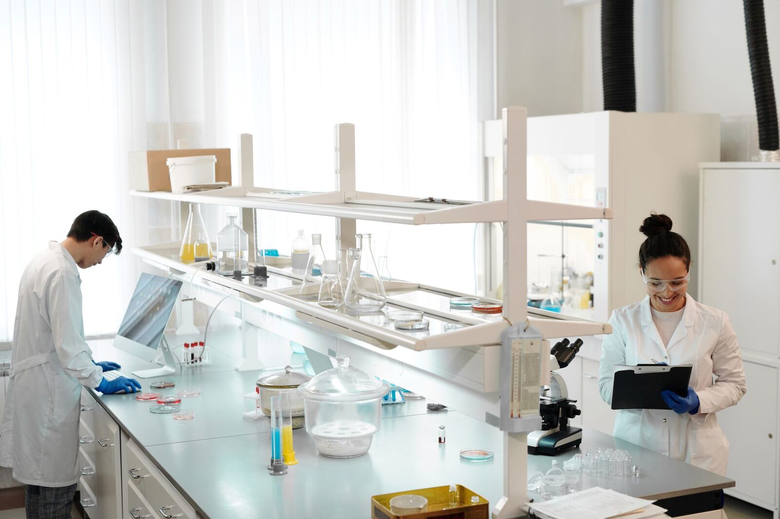 Two researchers at a laboratory bench, one entering data at a computer and one recording readings on a clipboard beside a microscope and rows of petri dishes.