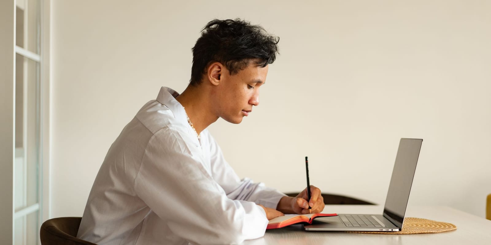 A student sitting at a table beside a window, writing notes in an open notebook next to an open laptop, the part of a nutrition degree that genuinely happens online