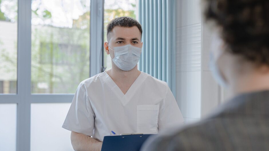 A clinician in white scrubs takes notes on a clipboard while sitting across from a patient in a hospital consulting room