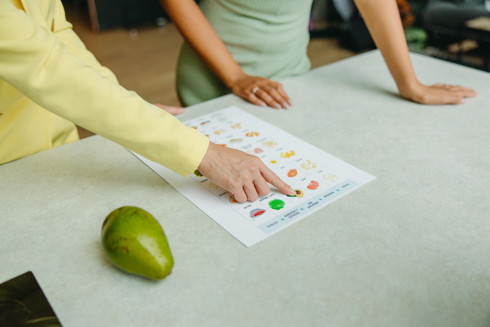 A dietitian points to a meal-composition chart laid out on a counter while a client stands across from her, the counselling side of a registered dietitian’s caseload.