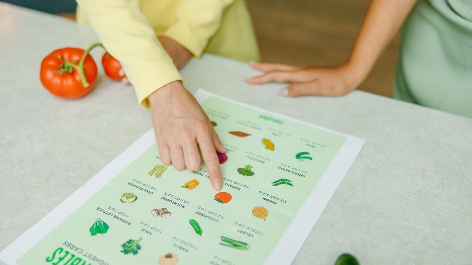 A nutrition professional points to an item on a printed vegetable reference chart while a client follows along across the table