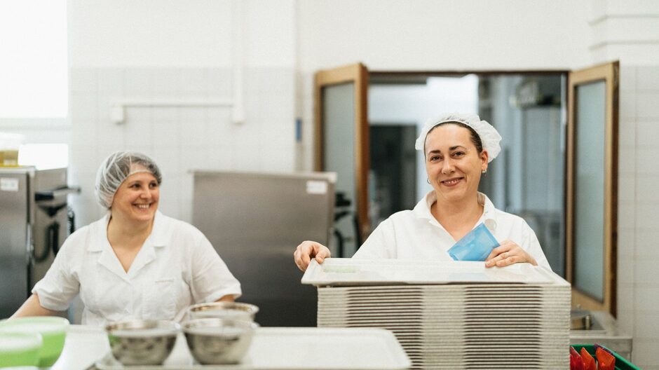 Two foodservice workers in white uniforms and hairnets stacking meal trays in an institutional kitchen