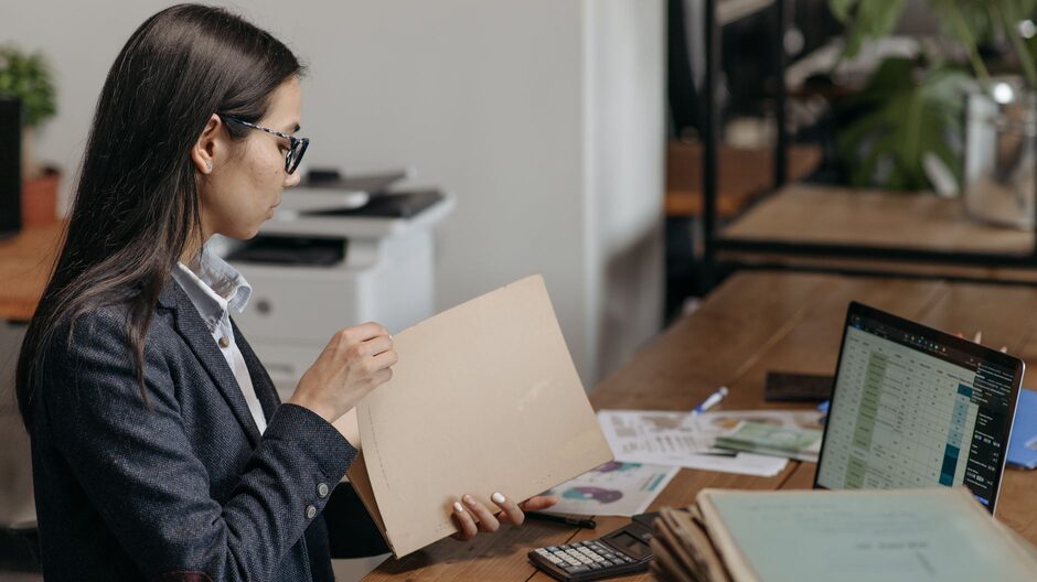 A professional at a desk reading through a case file, with stacked folders beside a laptop showing a spreadsheet