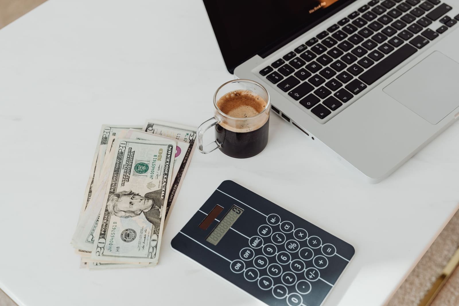 A white desk seen from above with an open laptop, a glass cup of black coffee, a fan of twenty-dollar bills and a handheld calculator