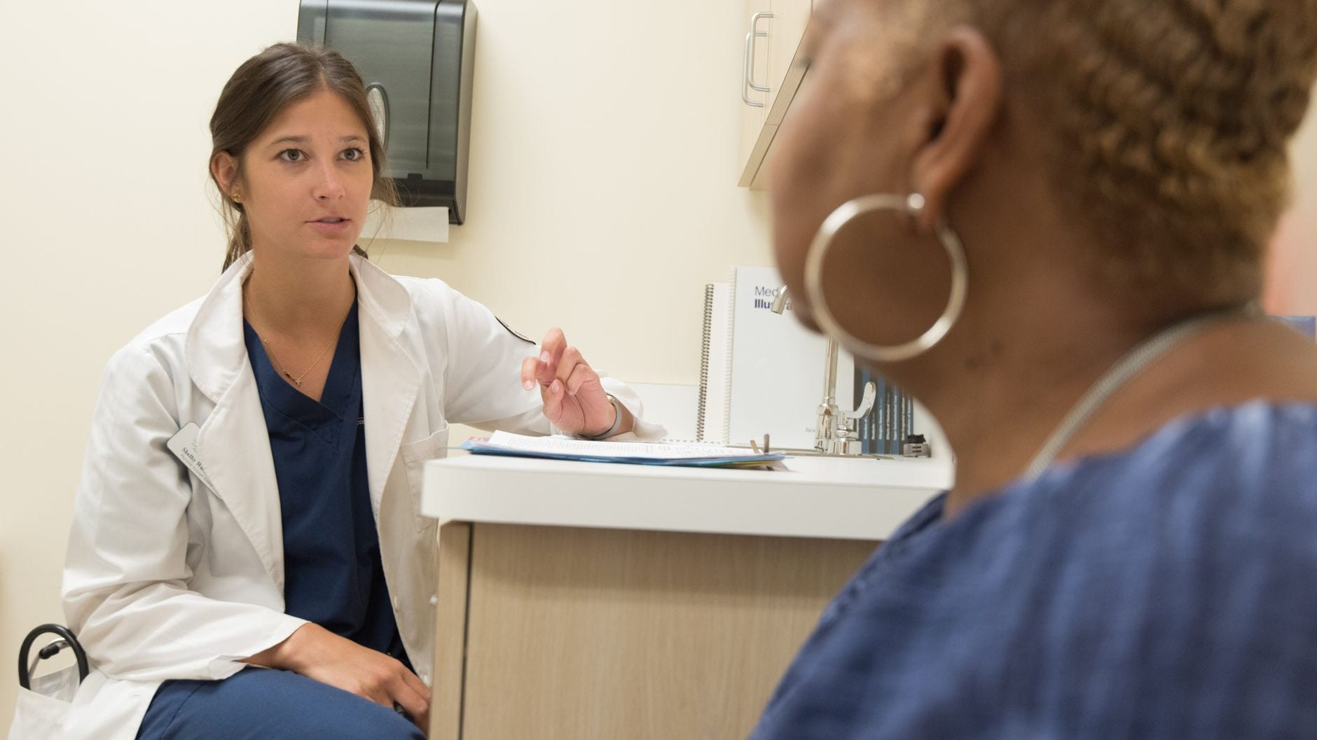 A clinician in a white coat and navy scrubs talking a seated patient through notes in a clinic exam room