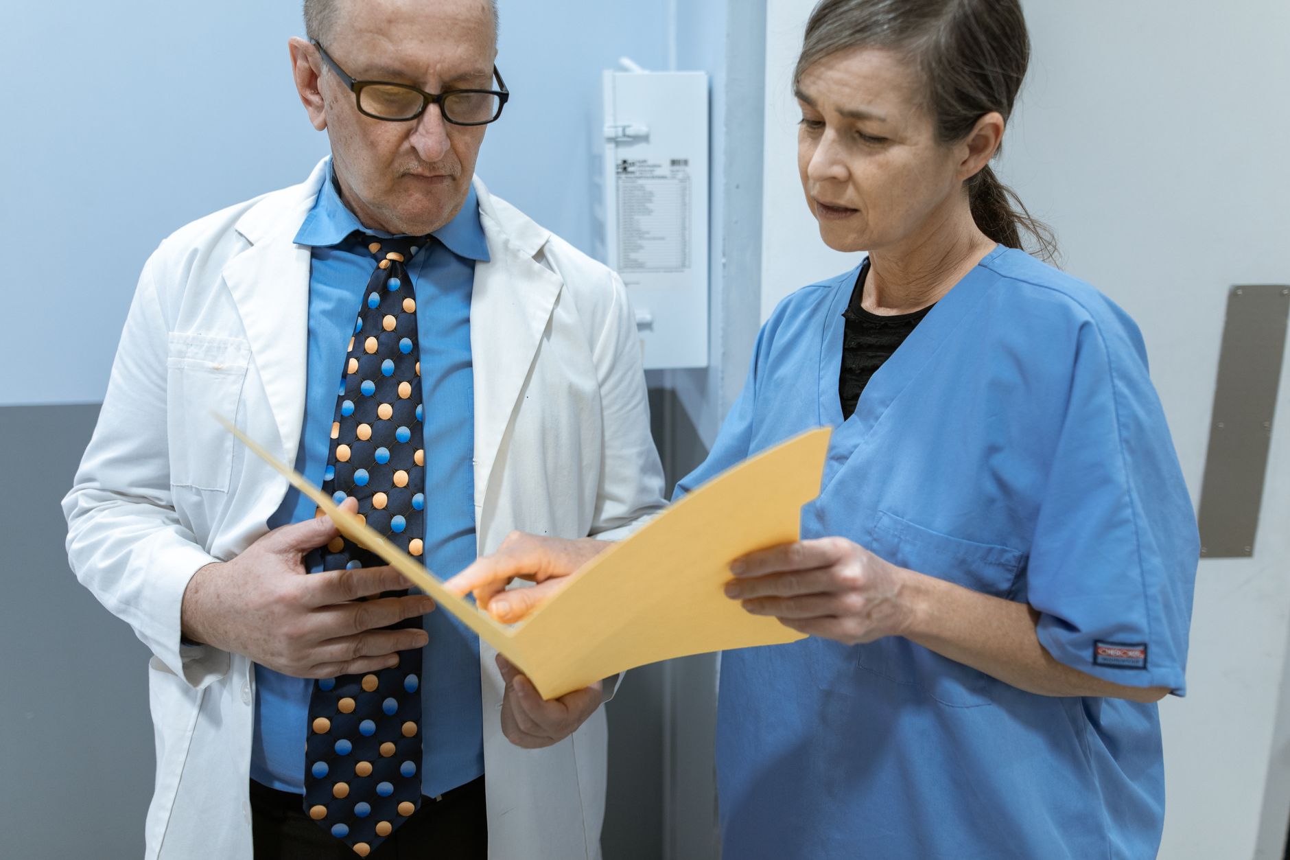 A clinician in a white coat and a colleague in blue scrubs stand together in a hospital corridor, reading a patient case file held open between them
