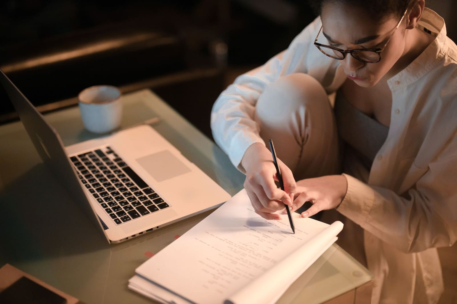 A woman in glasses sitting on the floor at a low table in the evening, marking up printed pages with a pencil beside an open laptop and a coffee cup