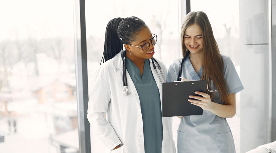 Two clinicians standing by a window, one in a white coat and one in scrubs, reading a patient chart together on a clipboard