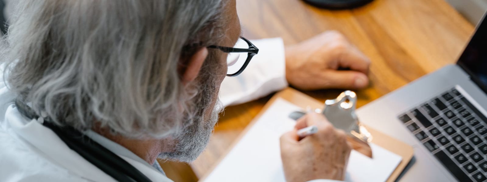 A clinician in a white coat with a stethoscope round the neck, writing notes on a clipboard at a desk beside an open laptop