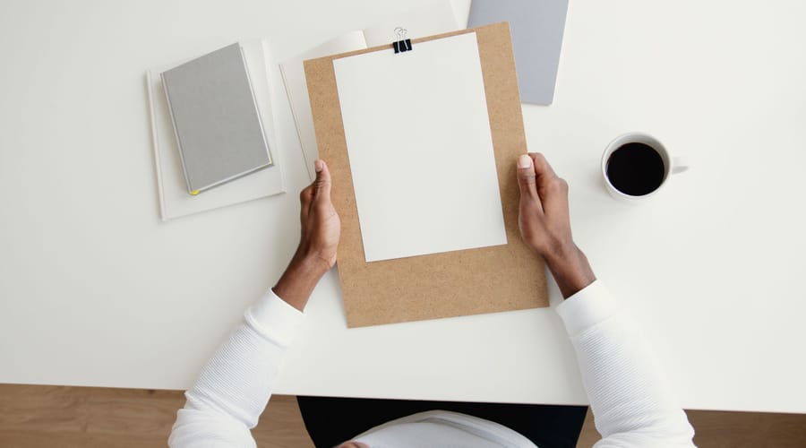 Overhead view of a person holding a clipboard with a blank scoring sheet clipped to it, beside a notebook and a cup of coffee